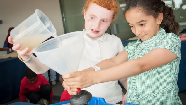 a student holds a funnel in outstretched arms while another student pours water in from a jug