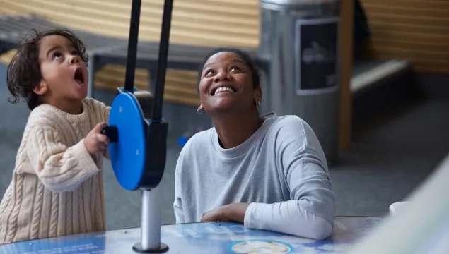 A woman looks up smiling. She is next to a young child with a look of amazement on their face. They are playing with a blue exhibit in We The Curious