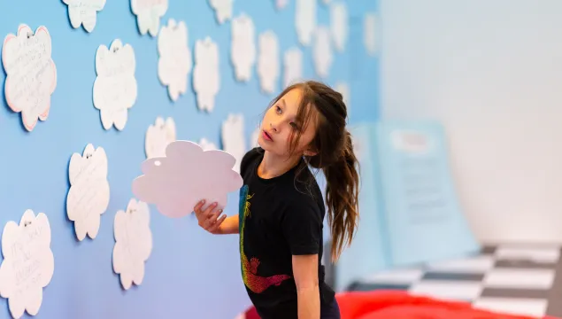 A child at an interactive learning exhibit, holding a cloud-shaped cutout next to a wall filled with similar cutouts.