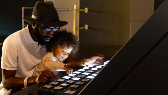 An Adult and a child are sat down playing with an exhibit that has light up buttons, and the child is pressing the buttons