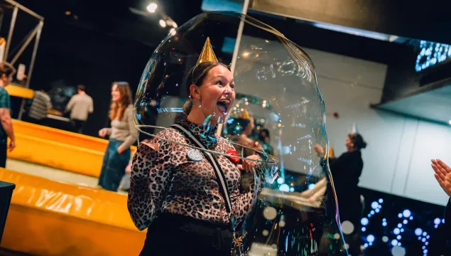 A person is in the middle of a science centre hold a giant bubble over their head.