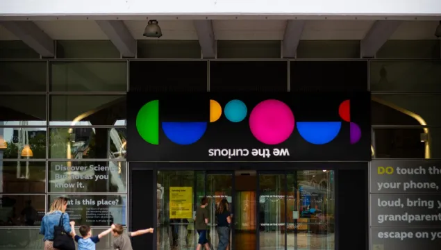 An image of the front of We The Curious, taken from the outside. Three visitors have their backs to the camera and are pointing up at the We The Curious logo