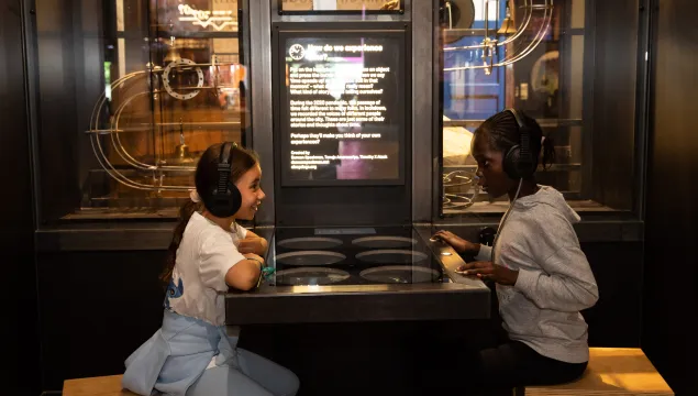 Two children are sat in a science museum opposite each other with a table in the middle of them. They are both wearing headphones 