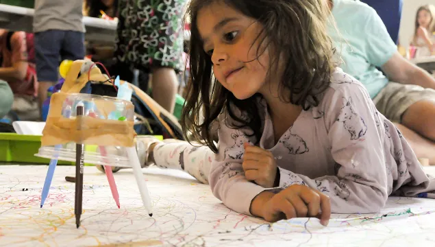 A small child is lying on the floor next to a creation made with a circuit and pens.