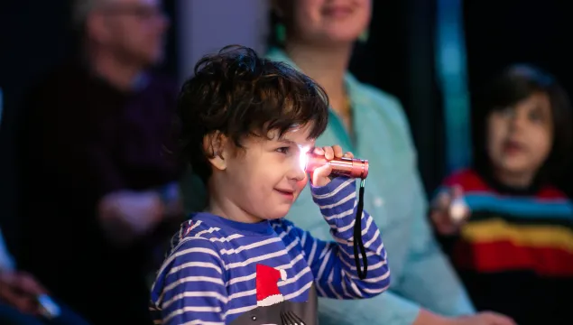 A small child is holding a torch up to it's eye, in a studio space.