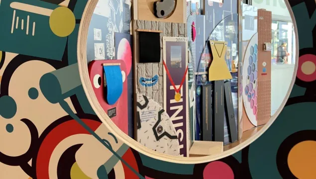 A round window in a colourful wall revealing an art display with geometric shapes, mirrors, and interactive elements in the foyer at We The Curious  