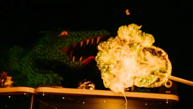 A dragon is in the background of the image with it's mouth open. To it's right in the foreground is a bright yellow flame. Taken from a demonstration from Fariytale SOS studio show at We The Curious