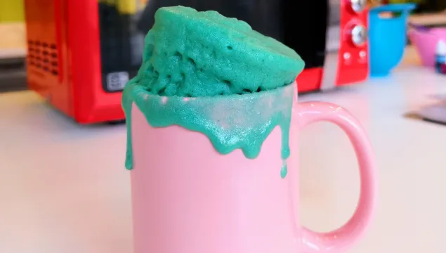 A pink mug is in front of a microwave with a bright blue mug cake spilling out of it onto the sides.