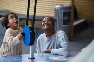 A woman looks up smiling. She is next to a young child with a look of amazement on their face. They are playing with a blue exhibit in We The Curious