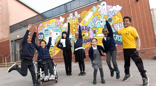 A cheerful group of children in school uniform in front of a bright colourful mural on a brick wall. They're all smiling, with arms in the air and some mid-jump