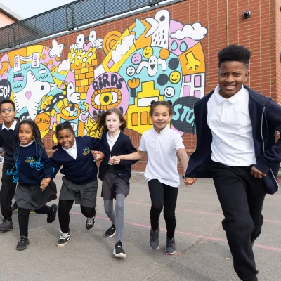 A group of school children in school uniform in front of a brightly coloured mural - running towards the camera. Credit Freia Turland