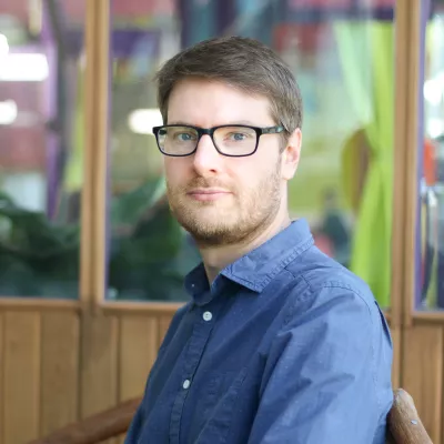 A portrait headshot of a person with short brown hair, glasses and a blue shirt, looking at the camera