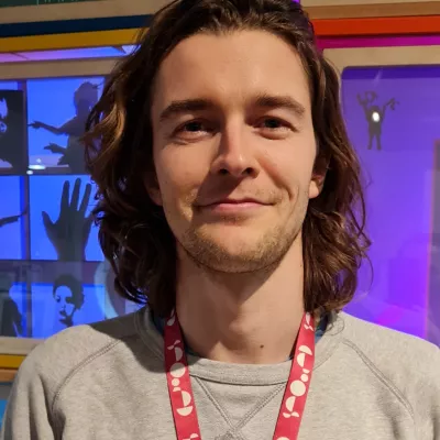 A portrait headshot of a smiling person with shoulder-length brown hair, a grey jumper and pink lanyard, looking directly at the camera. 