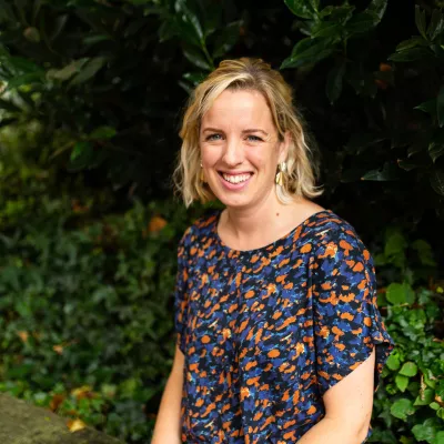 Person with shoulder length wavy blond hair smiling and looking into the camera in front of green plants