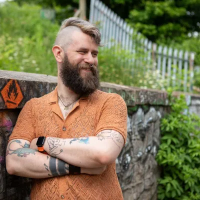 A person with brown beard, hair shaved on the side and long on top - smiling. They're stood with arms crossed, leaning against a wall, with green foliage behind 