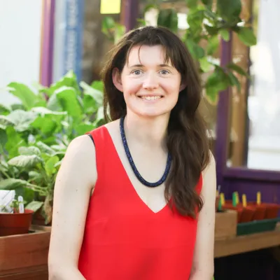 A person with long brown hair, smiling. They're sat down, wearing a red sleeveless top, with leafy foliage behind them. 