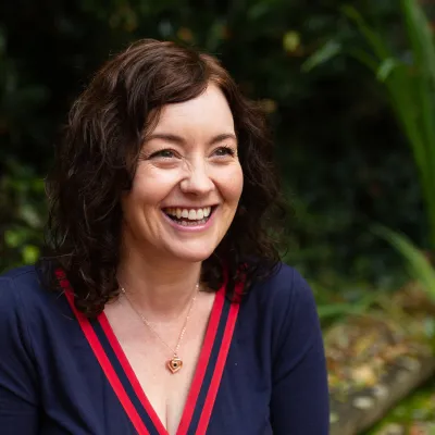 A person smiling with medium length brown hair, they are wearing a navy and red top and are sitting in front of some greenery.