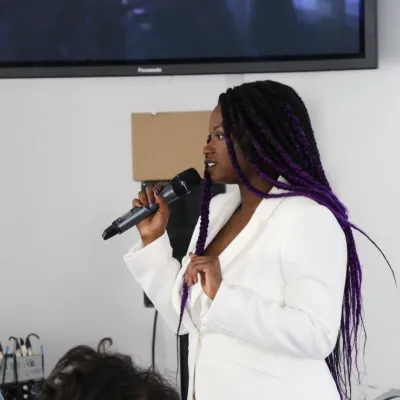 A profile view of a person speaking into a microphone, they have long dark hair, are wearing a white suit jacket and are in front of a plain wall with a mounted TV.
