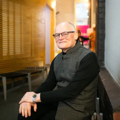 A person smiling with very short grey hair and glasses, they are wearing a black and grey jacket, and are sitting in front of a We The Curious exhibit.