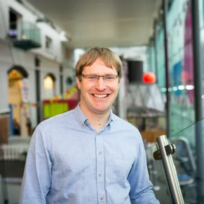 A person smiling with short light brown hair and glasses, they are wearing a blue shirt, and are standing in front of a We The Curious exhibit.