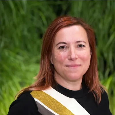 A person smiling with long reddish brown hair, they are wearing a black top with a white, yellow, and grey stripe and are in front of some greenery.