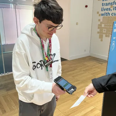 A young person wearing a white hoody holding a ticket scanning device, scanning a ticket for someone entering We The Curious 