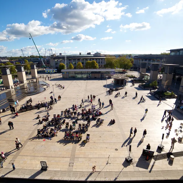 A large paved, open space with a crowd of people in the middle. To the left is a water feature. To the right is the top of The Planetarium which looks like a large mirrored, silver ball. 