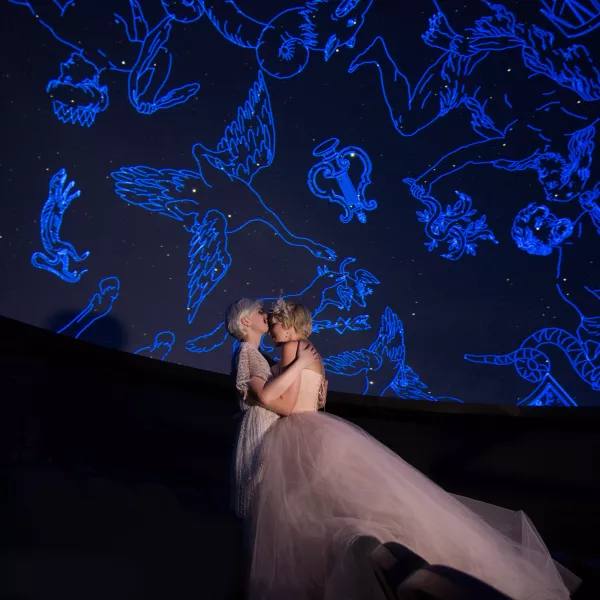 Two women in wedding dresses embracing each other standing inside the Planetarium with constellations projected onto the screen in the background