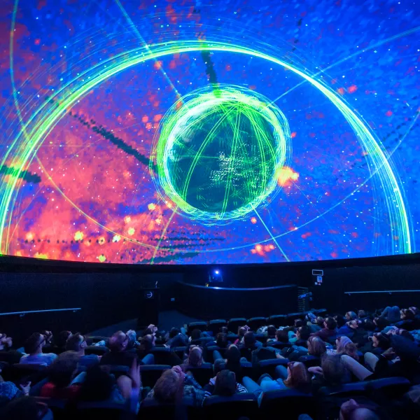 A shot of people sat in the Planetarium, a cinema-like experience. On the domed-shaped screen there's a visual of a colourful nebula.