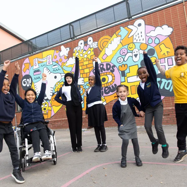 A cheerful group of children in school uniform in front of a bright colourful mural on a brick wall. They're all smiling, with arms in the air and some mid-jump