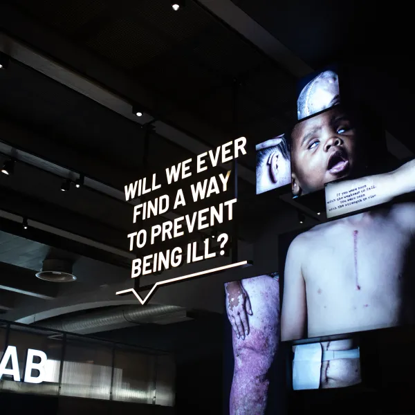 A lit up, hanging sign in the venue saying, 'Will we ever find a way to prevent being ill?'. Next to it are large backlit photos depicting different body parts