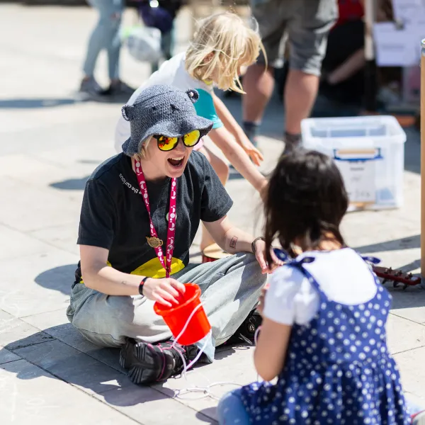 A Live Science Team member sat down outside in the sun, cross legged towards the camera. She has a small red bucket in her hand and is wearing sunglasses and a sun hat. She is smiling enthusiastically opposite a young girl who is wearing a blue polka dot dress facing away from the camera.