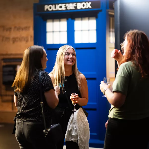 A group of three women standing amoung the exhibits talking and laughing with drinks in-hand