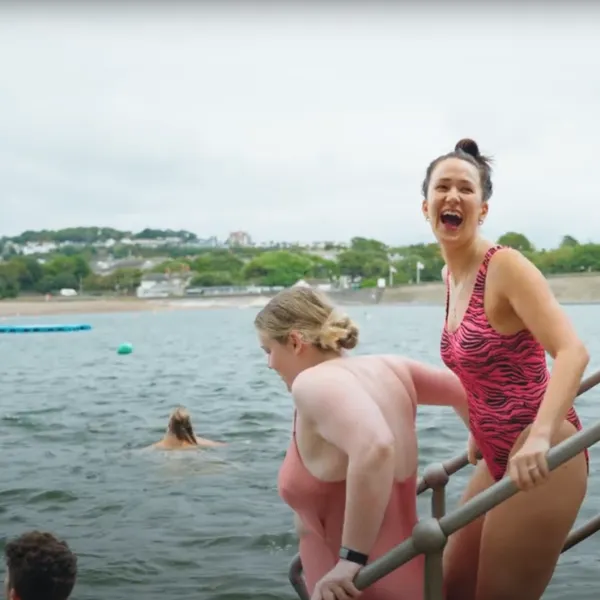 Three women in bathing costumes walking down steps into a body of water. The girl in the middle has her head turned towards the camera and is laughing.