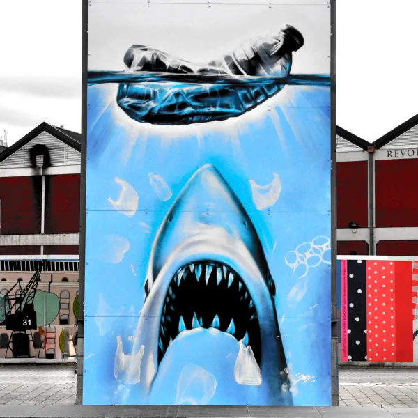 A public artwork showing a shark rising towards the surface towards a large plastic bottle, with red text reading 'Cause'