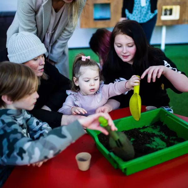 A family with a young child at the centre, sat around a red table with a tray of soil in the middle.