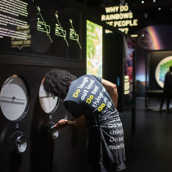 A member of We The Curious staff turns a handle on an exhibit. The question 'why do rainbows make people happy' is visible in the background. 