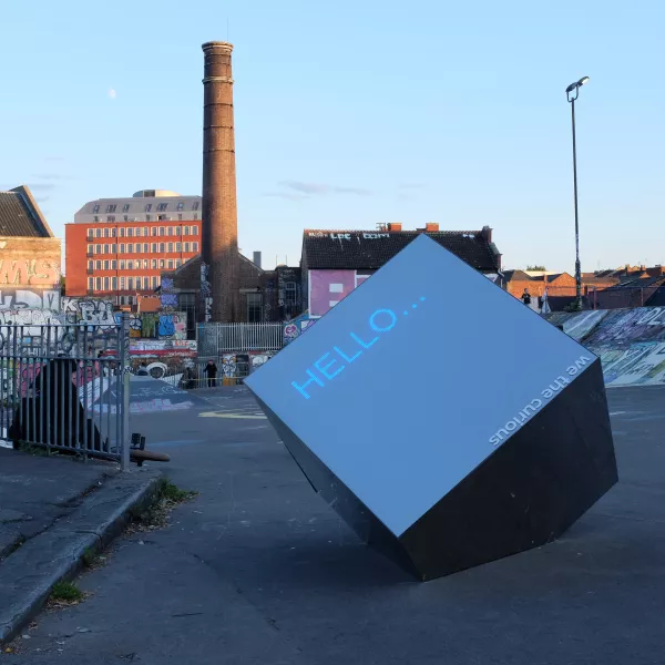 A large shiny metal cube reading 'hello...' with an urban skatepark landscape