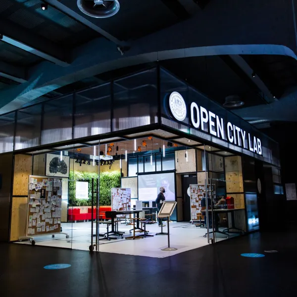 An brightly lit open space with tables, boards and activities with the name 'Open City Lab' space illuminated above 