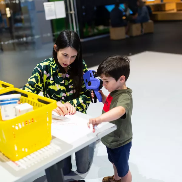 An adult and young child play with a hand-held pricing machine in the Open City Lab at We The Curious. There are yellow shopping baskets with some food containers in them on the table in front of them.