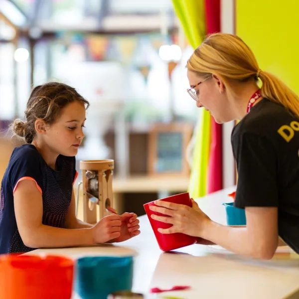 An adult and child are together in a kitchen space with brightly coloured bowls on a white surface. The adult is on the right, holding a red pot in their hands, which both the adult and child are looking into. The adult is wearing a grey ‘We The Curious’ uniform t-shirt and pink lanyard.