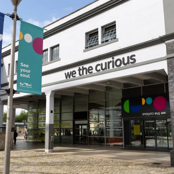 A view of the We The Curious building showing the main doors with the name and logo above. There is a colourful flag in the foreground that reads 'see your soul'.