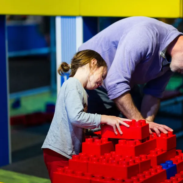 An adult and a child build a structure with large red bricks. 