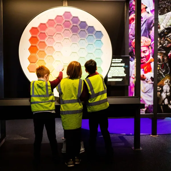 A group of children in high vis jackets look at the colour wheel at We The Curious, with their backs to the camera.