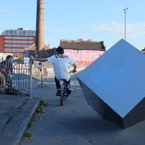 A young person on a bike has his back to the camera. They are in a skate park with industrial buildings in the background. Next to the young person is a large cube. On it are the words 'Will we ever travel at the speed of light? Phoebe, aged 11'