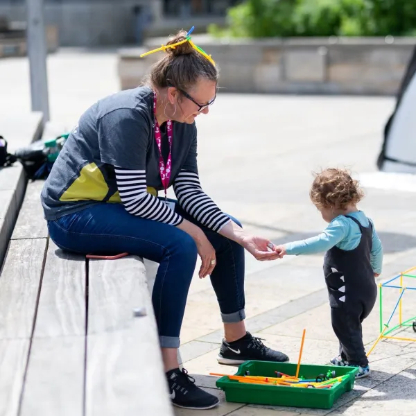 An adult and toddler playing with colourful straws together. The toddler is putting something in the adult's hand, they're outside in a stone paved space, with the adult sat on a wooden bench. 