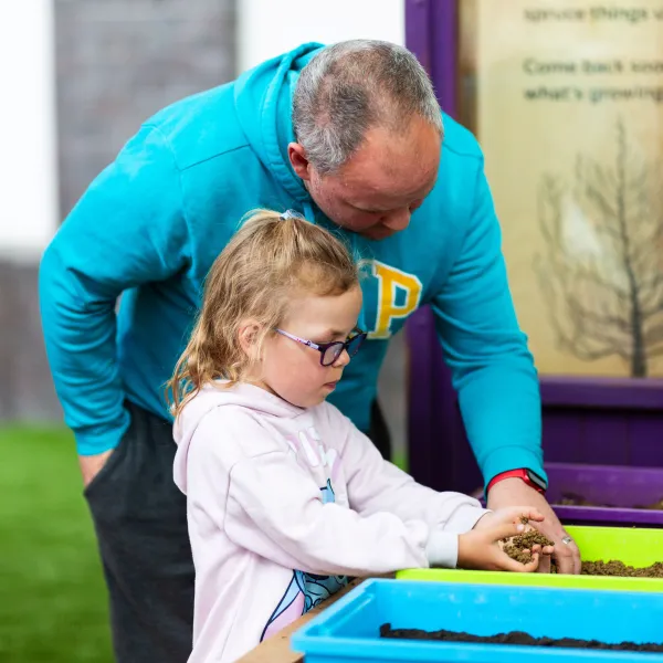 An adult and a child are in a greenhouse playing with soil in plastic containers.