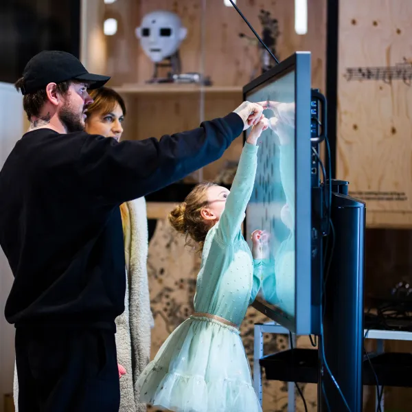 Two adults are helping a child who is reaching up and pointing at something on a screen.
