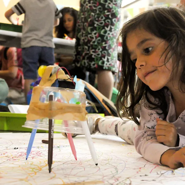 A small child is lying on the floor next to a creation made with a circuit and pens.