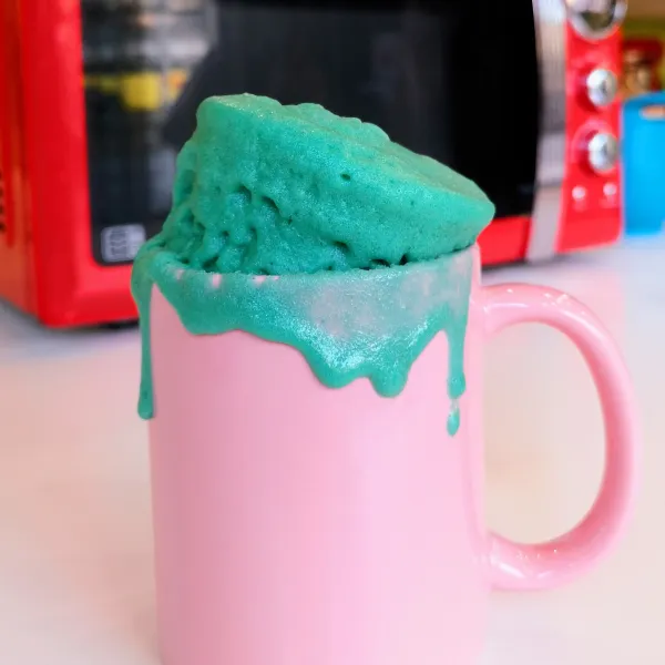 A pink mug is in front of a microwave with a bright blue mug cake spilling out of it onto the sides.